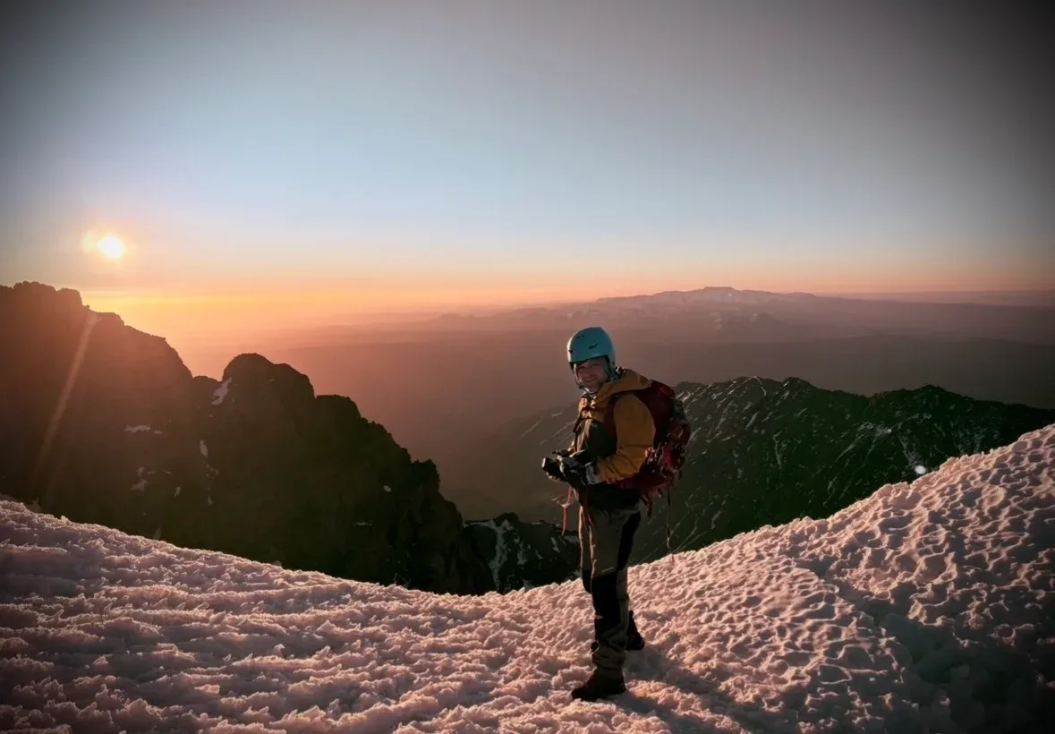 A helmeted mountaineer on a snowy ridge at sunrise, with mountains in the distance