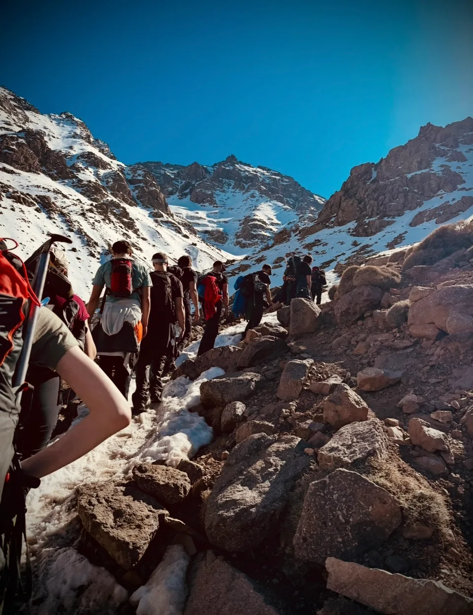 A group with rucksacks walking up a rocky path between snow-covered mountain slopes