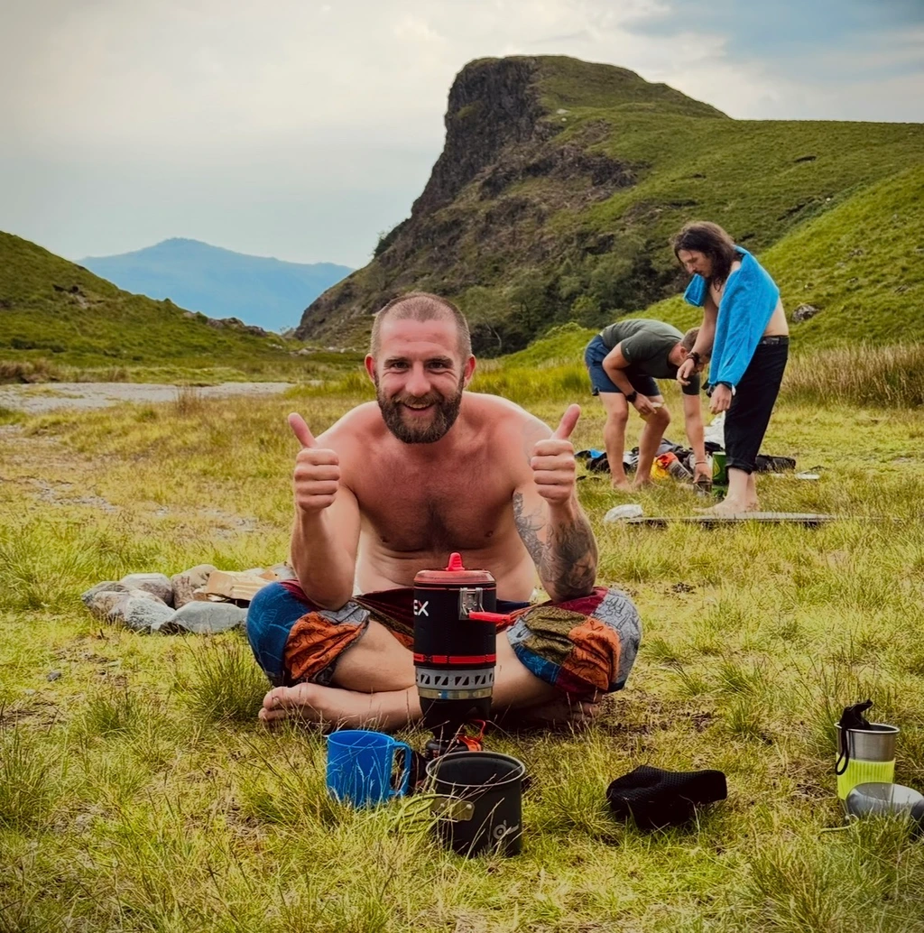 Dom sitting beside a mountain tarn with his camping stove, smiling and giving two thumbs up.