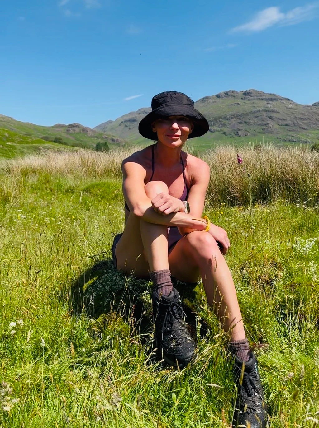 Keeley sitting in a sunny mountain meadow, wearing a walking hat and hiking boots.