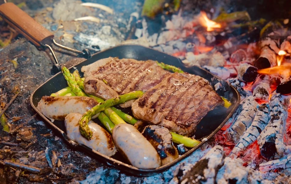 Steak, sausages and asparagus cooking in a pan over a campfire