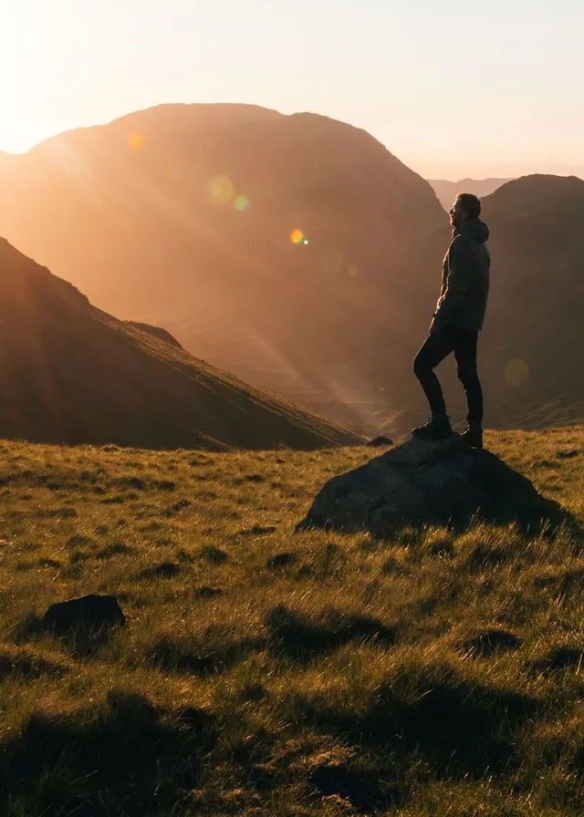 A walker standing confidently among sunlit Cumbrian fells, looking out across the mountains